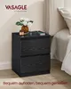 A black nightstand with two drawers is placed next to a bed. A vase with white flowers, a candle, and a smartphone are on top of the nightstand. Produktbild-Vorschau