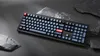 A black mechanical keyboard with gray and red keys is placed on a dark gray surface, surrounded by gray rocks. Produktbild-Vorschau
