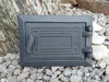A cast iron revision door for a chimney or oven is placed on a bed of gravel and rocks, with a large rock formation in the background. Náhled obrázku produktu