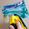 A hand is holding a yellow and black tool over a colorful artwork. Náhľad obrázku produktu