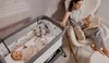 A woman is sitting in a rocking chair reading a book next to a baby crib with a baby inside. Produktbild-Vorschau