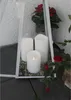 Three LED candles are placed inside a white lantern with gravel at the bottom. Red flowers are visible in the background and to the right of the lantern. Produktbild-Vorschau
