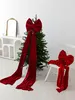 A large red velvet bow is placed on top of a decorated Christmas tree, with another bow on a chair in the foreground. Podgląd zdjęcia produktu