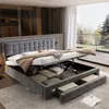A modern bedroom with a large bed featuring a hydraulic storage system and drawers, showcasing its functionality. The room has large windows with a view of trees and greenery. Produktbild-Vorschau