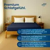 A bed with navy blue fitted sheets and white pillows, set in a bedroom with wooden furniture and a lamp. Produktbild-Vorschau