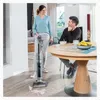 A woman is using a floor cleaner while a man sits at a table nearby. Produktbild-Vorschau