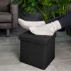 A person's feet in socks resting on a black ottoman in a living room setting. Náhľad obrázku produktu