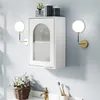A white bathroom cabinet with a frosted glass door is mounted on a light gray wall. Two gold and white sconces flank the cabinet, and a basket of white flowers sits on the counter below. Produktbild-Vorschau