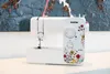 A white Brother sewing machine with floral decorations is placed on a white surface, with a textured artwork in the background. Produktbild-Vorschau