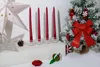 Four red spiral candles are displayed on a white surface, with a small pinecone and a decorative star in the background. A small, flocked Christmas tree with red ornaments and a large red bow is to the right. Náhled obrázku produktu