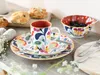 A colorful floral patterned dinnerware set with a mug, plates, and a bowl filled with blueberries, set on a wooden table with pastries in the background. Náhľad obrázku produktu