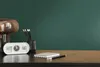 A desk with a clock, notebook, pencils, and a lamp against a dark green wall. Produktbild-Vorschau