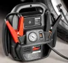 A black car jump starter with red jumper cable clamps is shown next to a car tire. Podgląd zdjęcia produktu