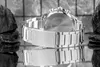 A close-up black and white shot of the back of a silver watch with a metal band, resting on a reflective surface with dark rocks on either side. Voir les images du produit