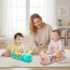 Zwei musikalische Krabbelraupen-Spielzeuge, eines türkis und eines rosa, auf einem Teppich mit einer lächelnden Frau und zwei spielenden Babys Anteprima dell’immagine prodotto