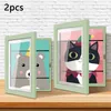 Two open picture frames displaying cartoon animal art are placed on a wooden surface against a white brick wall. Produktbild-Vorschau