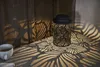A black metal lantern with a leaf pattern casts intricate shadows on a wooden surface and a concrete table next to a white cup of coffee. Produktbild-Vorschau
