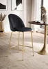 A black velvet bar stool with gold legs is placed next to a small round table in a well-lit room with a window and curtains. Voir les images du produit