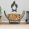 A modern black metal firewood rack filled with logs, placed on a wooden floor in front of a light gray wall with a graphic of a campfire. A plant in a basket is to the left, and a fireplace tool set is to the right. Podgląd zdjęcia produktu
