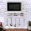 A microwave oven is placed on a white shelf mounted on a white brick wall. Below the shelf, kitchen utensils are hanging from hooks. A bottle of sauce is on the counter to the left. Podgląd zdjęcia produktu