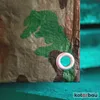 A close-up view of a camouflage tarp with a grommet, resting on a green surface. Produktbild-Vorschau