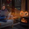 A woman is sitting on a couch using a laptop, with a wood-burning stove and a fan in the background. Produktbild-Vorschau