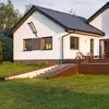 A modern white house with a dark roof, featuring a solar street lamp mounted on the wall. The house has large windows and a wooden deck with lounge chairs. A grassy lawn and a set of stairs lead up to the house. Produktbild-Vorschau
