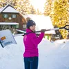 A woman is holding a snow shovel over her shoulder in a snowy environment with a house in the background. Produktbild-Vorschau