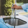 A person's hand is holding a pull-down faucet head over an outdoor kitchen sink, with water spraying into the sink. Náhled obrázku produktu