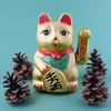 A golden Maneki-neko cat figurine is displayed on a teal background, flanked by pinecones. Produktbild-Vorschau