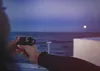 A person holding a coffee cup while looking out at the ocean at dusk with the moon visible. Produktbild-Vorschau
