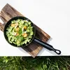 A miniature black frying pan filled with chopped green vegetables, sliced orange carrots, and small white flowers, placed on a piece of weathered wood. Produktbild-Vorschau