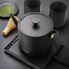 A black electric kettle with gold accents sits on a black heating base, next to a matcha whisk, a small plate with green powder, and two dark cups. Produktbild-Vorschau