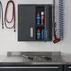 A dark gray metal cabinet with sliding doors is mounted on a white tiled wall above a stainless steel workbench. The cabinet contains various automotive fluids and cleaning supplies. Tools are laid out on the workbench. Voir les images du produit