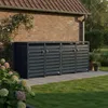 Four dark gray trash bins are lined up against a brick wall in a garden setting. There are plants and flowers in the foreground and background. Produktbild-Vorschau