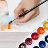 A hand is painting on a watercolor paper with a brush, next to a palette of watercolors. Podgląd zdjęcia produktu