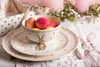 Easter table setting with decorated eggs in a bowl on plates, with decorative elements in the background. Náhled obrázku produktu