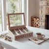 A wooden cutlery box with a glass lid sits on a white table next to a cup of tea and some dried herbs. A stone fireplace with stacked firewood is visible in the background. Podgląd zdjęcia produktu