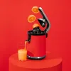 A red electric juicer is shown in action, with oranges being juiced and orange juice filling a glass. The background is a solid red. Podgląd zdjęcia produktu