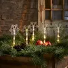 Snowflake lights are placed in a pine garland with pinecones and apples, set against a brick wall and a wooden door. Podgląd zdjęcia produktu