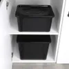 Two black plastic storage containers with lids are stacked on shelves inside a white cabinet. Voir les images du produit