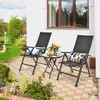 Two black folding chairs and a small round table are set up on a patio with flowers and greenery in the background. Náhľad obrázku produktu