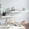 Two white floating shelves mounted on a light blue wall, decorated with various items including bottles, a box, a decorative cube, books, and artwork. A desk with more items is in the foreground. Náhľad obrázku produktu