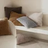 Several fluffy pillows in various neutral colors are arranged on a shelf and on the floor, with a portion of a cream-colored armchair visible in the foreground. Produktbild-Vorschau