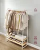 A white metal garment rack with two wooden shelves, filled with clothing, hats, shoes, and towels. The background is a light gray wall and a wooden floor. Náhled obrázku produktu
