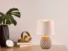 A table lamp with a textured ceramic base and a fabric shade sits on a wooden desk next to a vase with a monstera leaf, a stack of books, a golden sculpture, and a clock. Anteprima dell’immagine prodotto