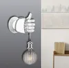 A white hand-shaped wall lamp with a lightbulb hanging from it, mounted on a gray wall. A piece of abstract art is visible on the right. Produktbild-Vorschau