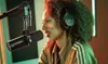 A woman with curly hair wearing headphones is speaking into a microphone in a recording studio. Náhľad obrázku produktu