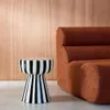 A black and white striped side table is placed next to a rust-colored sofa in a room with a wood-paneled wall. Produktbild-Vorschau