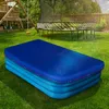An inflatable pool covered with a blue tarp sits on a grassy lawn in a backyard. A wooden playground structure and a trampoline are visible in the background. Produktbild-Vorschau